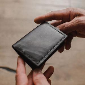 a black leather bifold wallet by Solid Leather being held by a pair of mans hands with a blurred background