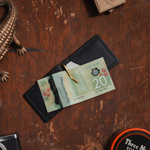 an open black full-grain leather money clip wallet showing $20 Canadian bill displayed on a wooden table surrounded by vintage props