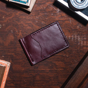 A rich burgundy leather cardholder wallet with visible stitching, placed on a dark wooden desk surrounded by vintage props including a camera, books, and a brass object.
