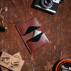A cognac brown leather card holder wallet with a geometric cutout design, shown in a styled flat-lay on a rustic wooden table alongside a camera, a lizard figurine, and other vintage props.