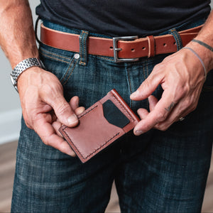 A man wearing a black t-shirt and blue jeans holds a slim cognac brown leather money clip card holder wallet in front of his waist. He is wearing a matching leather belt