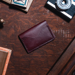 A dark burgundy leather card holder wallet with a stitched envelope-style flap, placed on a dark wooden tabletop surrounded by vintage props including a camera, notebook, and metal object.