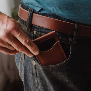 A close-up of a person wearing dark blue jeans and a cognac brown leather belt, with their hand pulling out a matching cognac brown leather card holder or wallet tucked into the jeans pocket.