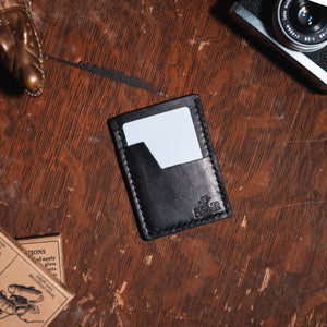 a black full-grain leather slim cardholder wallet by Solid Leather sits on a dark wooden table with vintage props. Handmade by Jesse Bateson in Peterborough Ontario Canada