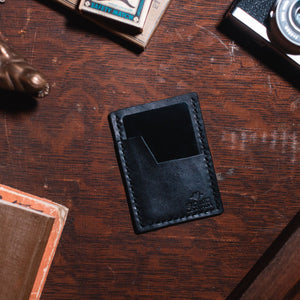 A black leather card holder wallet with a slanted card pocket, shown from above on a dark wooden desk surrounded by vintage props including a camera, a small figurine, and a book.