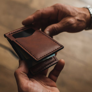 A close-up of a person's hands holding an open cognac brown leather card holder wallet, showing its interior compartments and card slots, with a blurred warm-toned background.