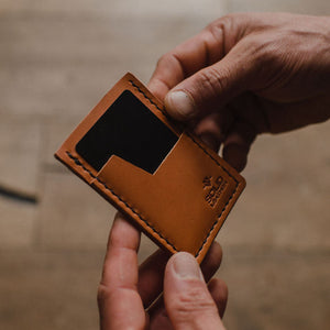 A close-up of a person's hands holding a small tan leather card holder minimalist wallet against a blurred wooden background. Handmade in Peterborough Ontario Canada.