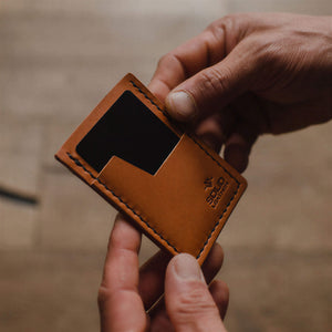 A close-up of a person's hands holding a small tan leather card holder minimalist wallet against a blurred wooden background. Handmade in Peterborough Ontario Canada.