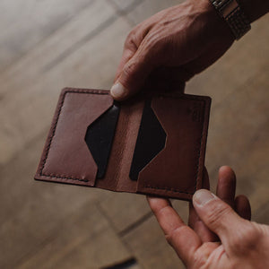 a pair of mans hands holding a cognac brown solid leather bifold cardholder wallet showing the inside card sleeves on a blurred background. Handmade by Jesse Bateson