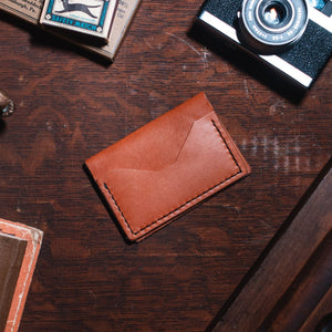 A tan leather bifold card holder wallet with a flap closure, placed on a dark wooden desk surrounded by vintage props including a camera, a brass object, and a book.