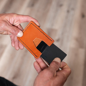 A close-up of a person's hands holding a full-grain tan leather card holder with a black card partially inserted, set against a blurred wooden floor background.