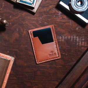 A tanleather card holder wallet with a black card slot, placed on a dark wooden desk surrounded by vintage props including a camera, a small figurine, and an old book.