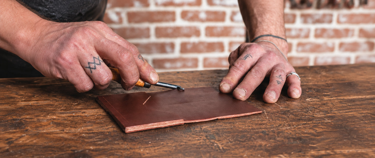 A close-up of Jesse Bateson's tattooed hands crafting a full-grain leather wallet on a wooden workbench at his Solid Leather studio in Peterborough Ontario Canada, with a blurred brick wall in the background.