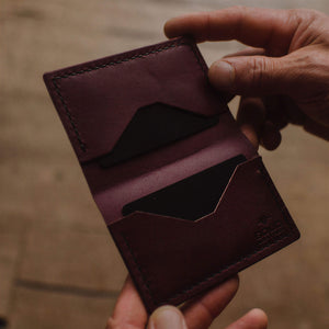 A close-up of a person's hands holding an open burgundy leather bifold cardholder wallet against a warm, blurred wooden background.