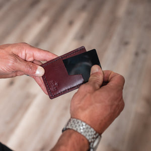 A close-up of a person's hands holding a burgundy leather card holder wallet with a black card inserted, set against a blurred wooden floor background.