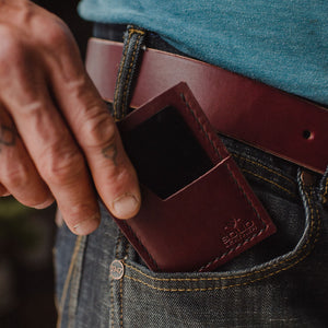 A close-up of a person's hand inserting a burgundy leather card holder into the front pocket of their blue jeans, tucked between a blue t-shirt and a brown leather belt.