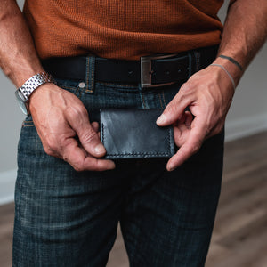 A close-up of a man's hands holding a slim black leather wallet bifold card holder at his waistband, wearing a rust-orange knit sweater, dark jeans, and a black belt in an indoor room with wood floors.