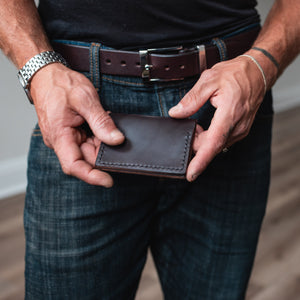 A close-up of a man's hands holding a dark brown leather bifold wallet, wearing blue jeans and a brown leather belt with a silver buckle.