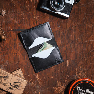 A black leather card holder wallet with a geometric cutout design, shown on a rustic wooden tabletop with vintage props including a camera, a lizard figurine, and a round tin.