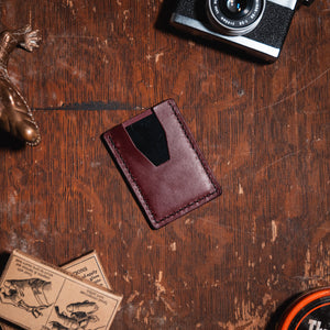 A burgundy leather card holder wallet with a black accent panel, displayed on a rustic wooden table alongside a bronze alligator figurine, a vintage camera, a stamp, and a round ink pad.