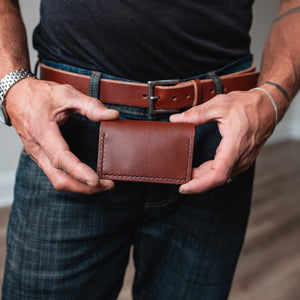 a full-grain leather bifold cardholder wallet in cognac brown leather is held by a pair of mans hands with a matching solid leather belt on a pair of blue jeans