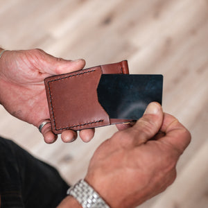 A close-up of a person's hands holding a cognac brown leather money clip card holder wallet with a black card inserted, set against a blurred wooden floor background.