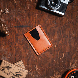 A tan leather card holder wallet with a black accent panel, displayed in a styled flat-lay on a rustic wooden table alongside a brass alligator figurine, a vintage camera, matchboxes, and a round tin.