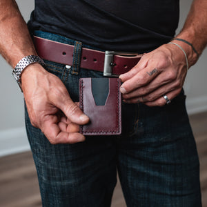 A close-up of a man's hands holding a burgundy leather card holder wallet, with his matching full grain burgundy leather belt and blue jeans in an indoor setting with a wood floor.
