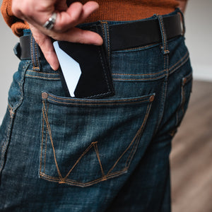 A close-up of a person wearing dark blue denim jeans with a black leather belt and a black card holder or wallet tucked into the back pocket, with a white card partially visible.