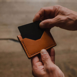 A close-up of a person's hands holding a Solid Leather slim, tan leather bifold card holder wallet with a black card partially inserted, set against a blurred wooden background.