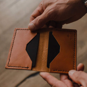 A close-up of a person holding an open tan leather bifold card holder wallet with black interior lining, showing its card slots and stitching, against a blurred wooden background. handmade in Peterborough Ontario by jesse bateson of Solid Leather