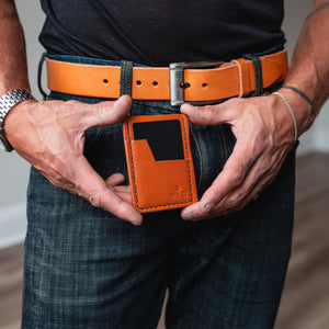 A close-up of a man wearing a dark t-shirt, blue jeans, a full-grain tan leather belt, and a matching tan leather card holder wallet, holding it open with both hands.
