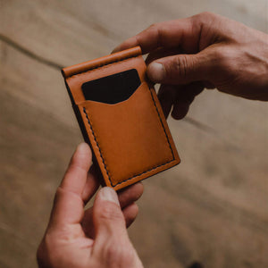 A close-up of a person's hands holding a slim, tan leather card holder wallet with a black card insert, set against a blurred wooden background. Handmade in Peterborough Ontario by jesse bateson of Solid Leather