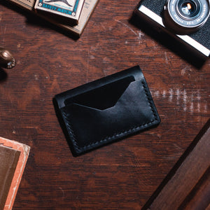 A minimalist black leather bifold card holder wallet with a single angled card slot, displayed on a dark wooden tabletop surrounded by vintage props including a camera, a small alligator figurine, and books.