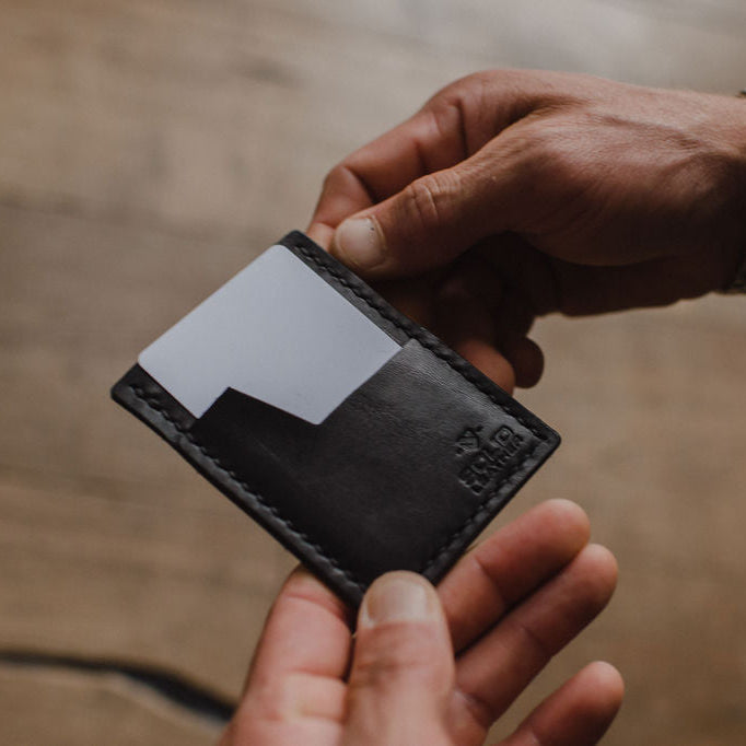 A close-up of a person's hands holding a black leather minimalist card holder wallet with a white card inserted, set against a warm, blurred wooden background.