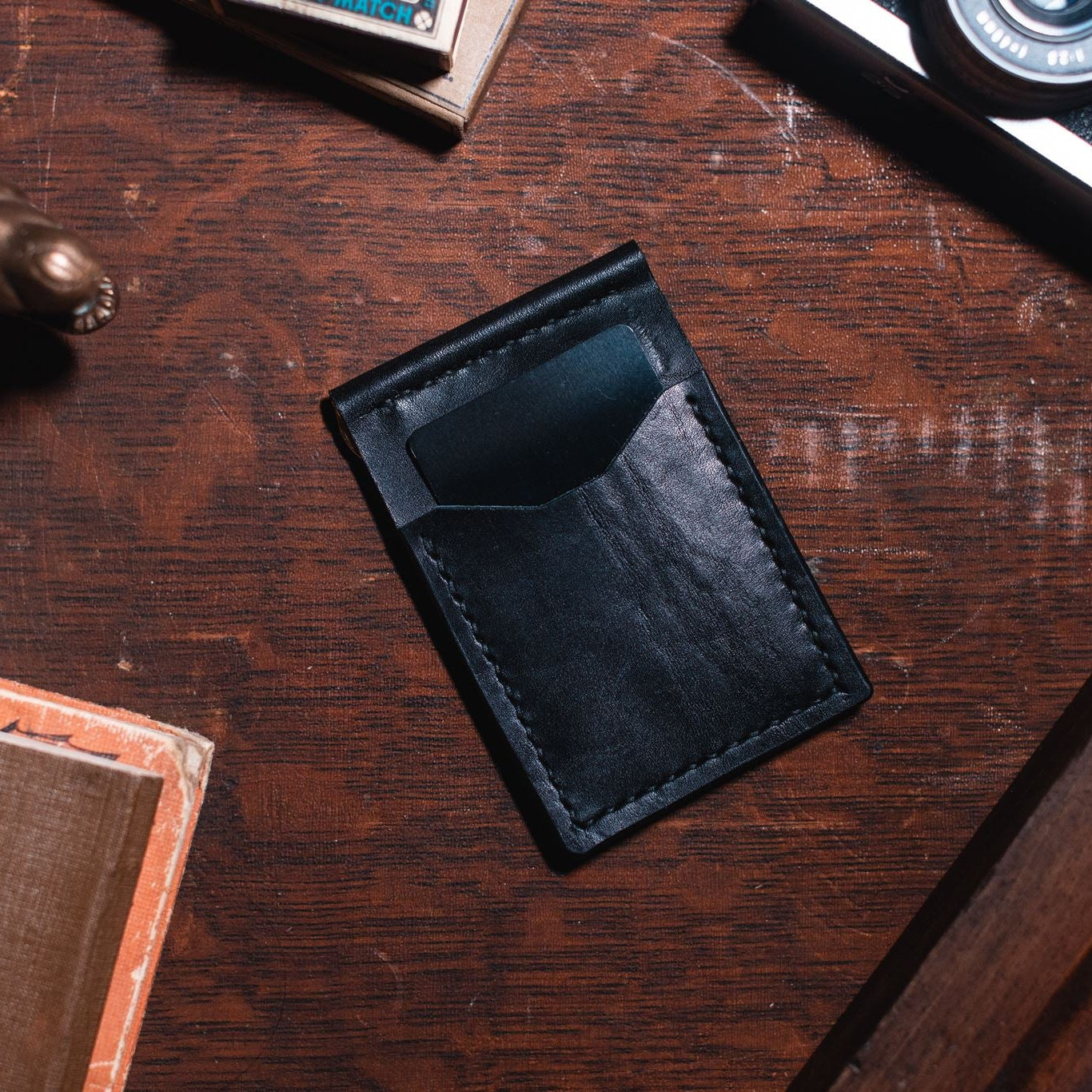 A black leather card holder wallet with visible stitching, placed on a dark brown wooden tabletop surrounded by vintage props including a camera, a brass figurine, and a book.