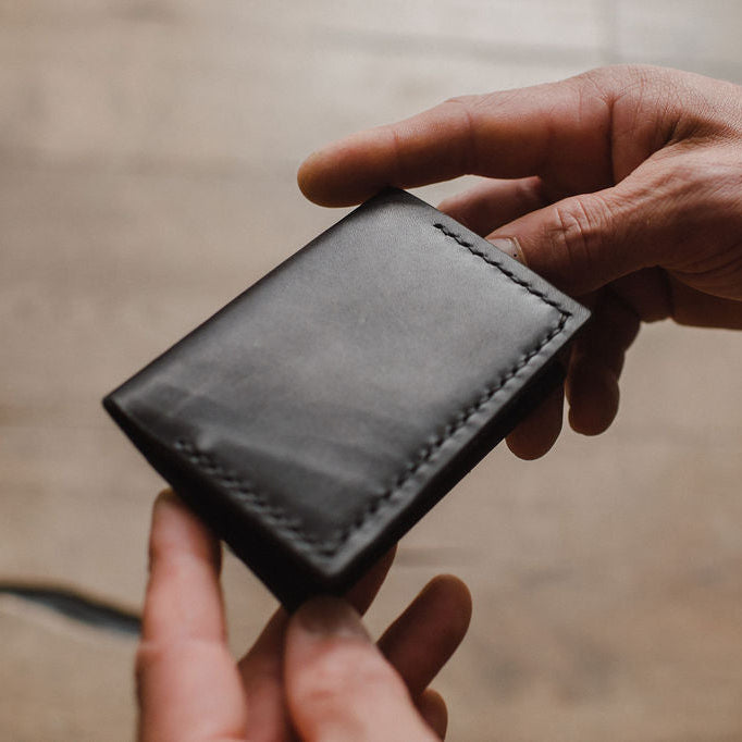 a black leather bifold wallet by Solid Leather being held by a pair of mans hands with a blurred background  