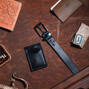 A black solid leather belt with a stainless steel buckle and a matching black leather card holder wallet, arranged on a dark wooden table alongside other vintage-style leather goods.