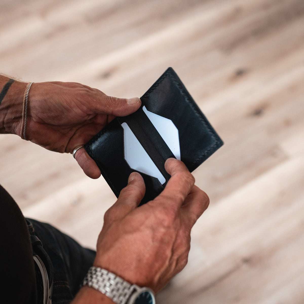 A close-up of a person's hands holding an open black leather wallet from solid Leather against a blurred light wood plank floor background.