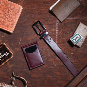 A styled flat-lay of a burgundy formal  leather belt with a stainless steel buckle and matching keeper, displayed on a dark wooden table alongside a matching leather wallet. 