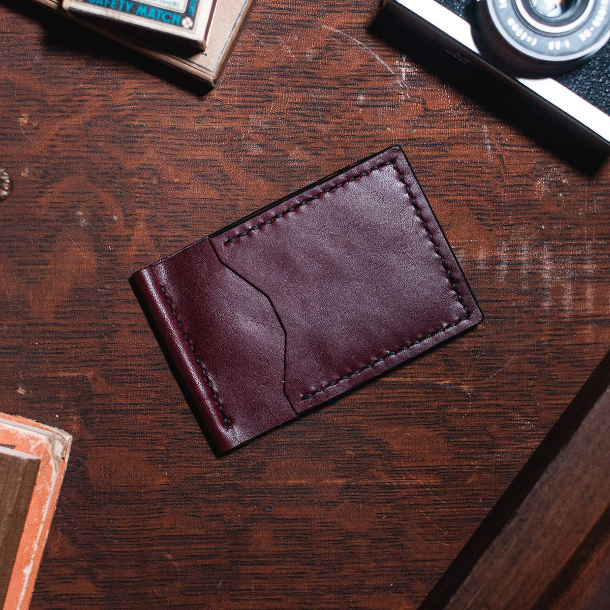 A rich burgundy leather cardholder wallet with visible stitching, placed on a dark wooden desk surrounded by vintage props including a camera, books, and a brass object.