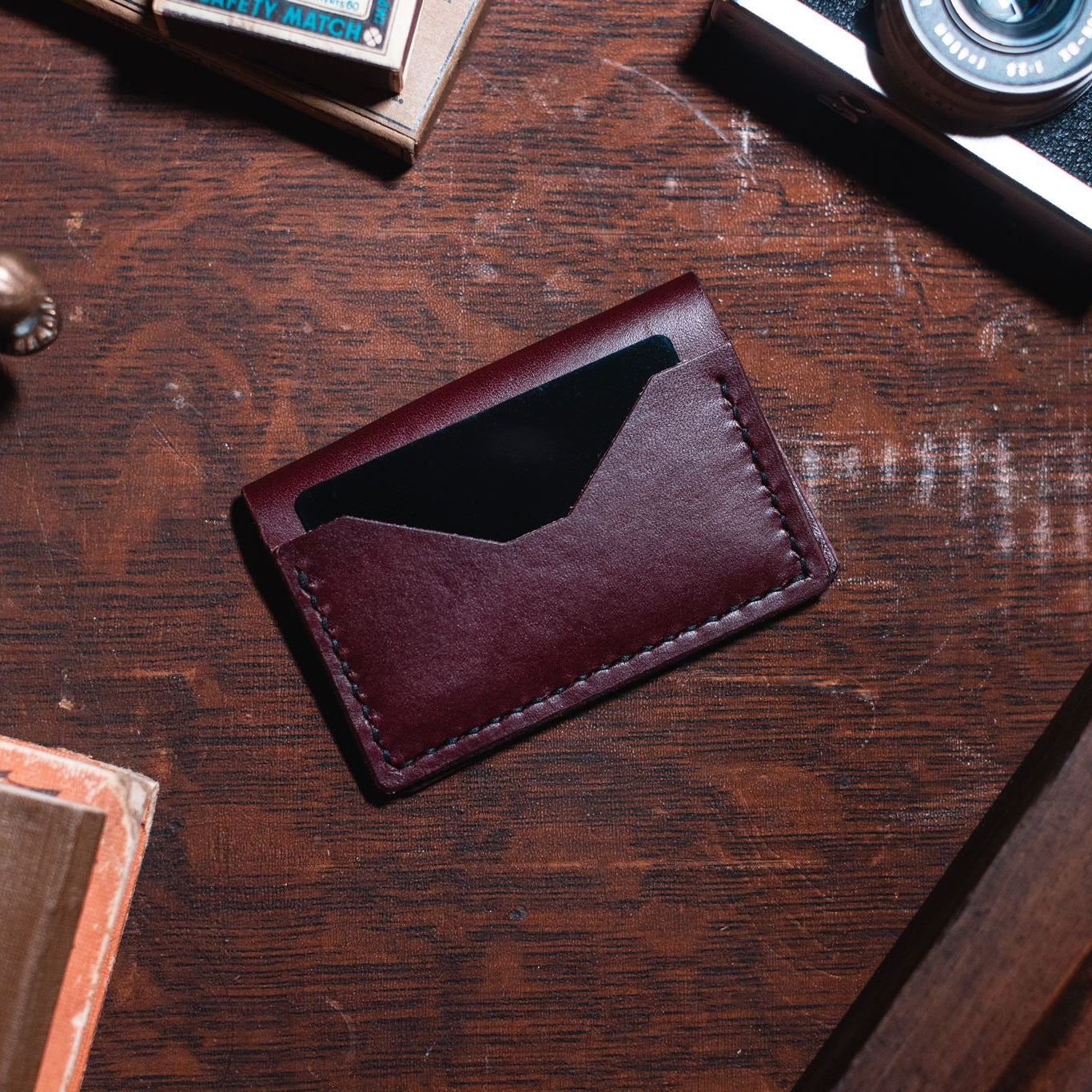 A burgundy leather bifold card holder wallet resting on a dark wooden desk surrounded by vintage props like a camera, a brass figurine, and old books. Handmade in Peterborough Ontario by Jesse Bateson of Solid Leather 