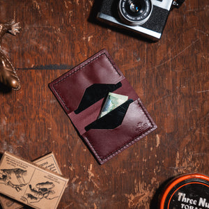 A dark burgundy leather cardholder wallet with a  geometric cutout design, shown on a rustic wooden tabletop with a camera, a lizard figurine, and other vintage-style props.