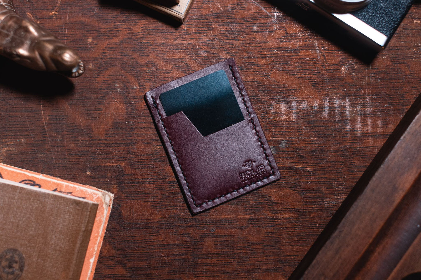 A burgundy leather minimalist card holder wallet with a teal card inserted, resting on a dark wooden desk with vintage props.