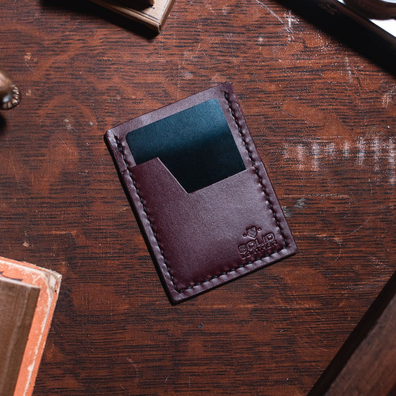 A burgundy leather minimalist card holder wallet with a teal card inserted, resting on a dark wooden desk with vintage props.