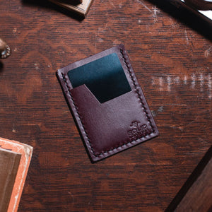 A burgundy leather minimalist card holder wallet with a teal card inserted, resting on a dark wooden desk with vintage props.