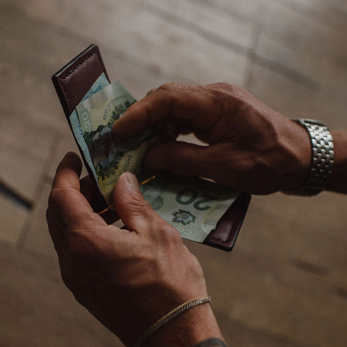 A close-up shot of a person's hands holding an open burgundy leather wallet and pulling out Canadian banknotes, with a wooden floor in the background.