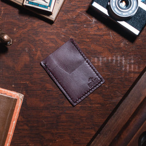 A burgundy leather card holder wallet with visible stitching and an embossed logo, placed on a dark wooden tabletop surrounded by vintage props including a camera, books, and a brass alligator figurine.