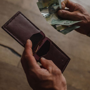 A close-up of a person's hands holding an open burgundy leather wallet built by Solid Leather in Peterborough Ontario and inserting Canadian momey, with a wooden floor in the background.