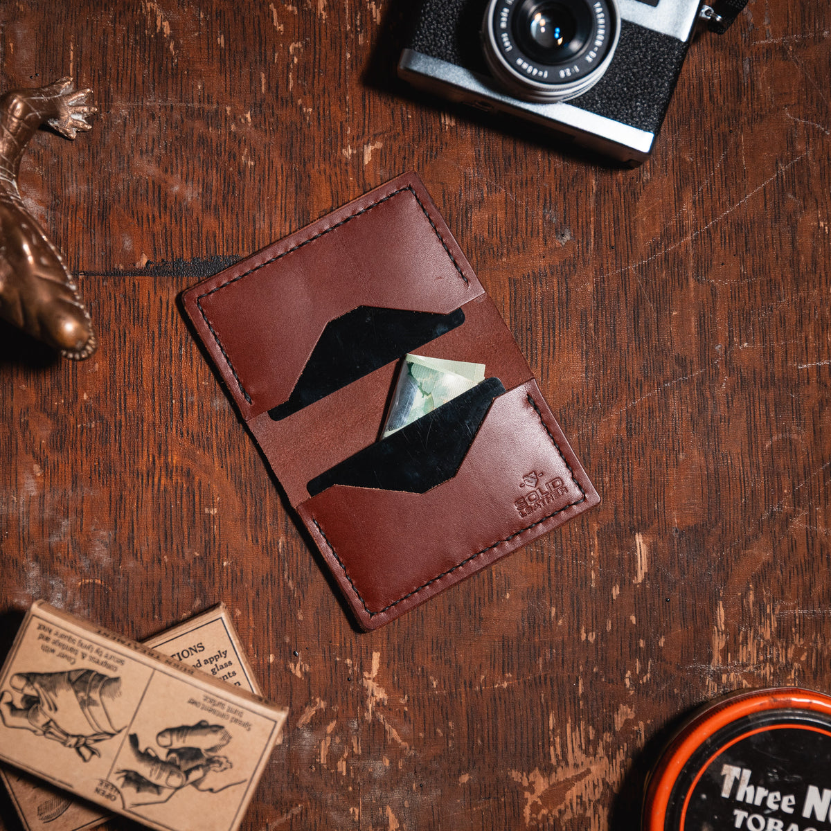 A cognac brown leather card holder wallet with a geometric cutout design, shown in a styled flat-lay on a rustic wooden table alongside a camera, a lizard figurine, and other vintage props.